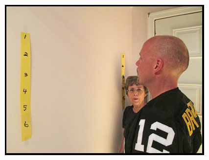 The yellow strip of paper is numbered 1-5 and is taped vertically on the wall about head-high to the left of where the man in the football jersey is standing.  An identical yellow, numbered strip of paper is on the wall to the right of the man, and Dona is watching the man as he looks on the wall between the strips of paper.